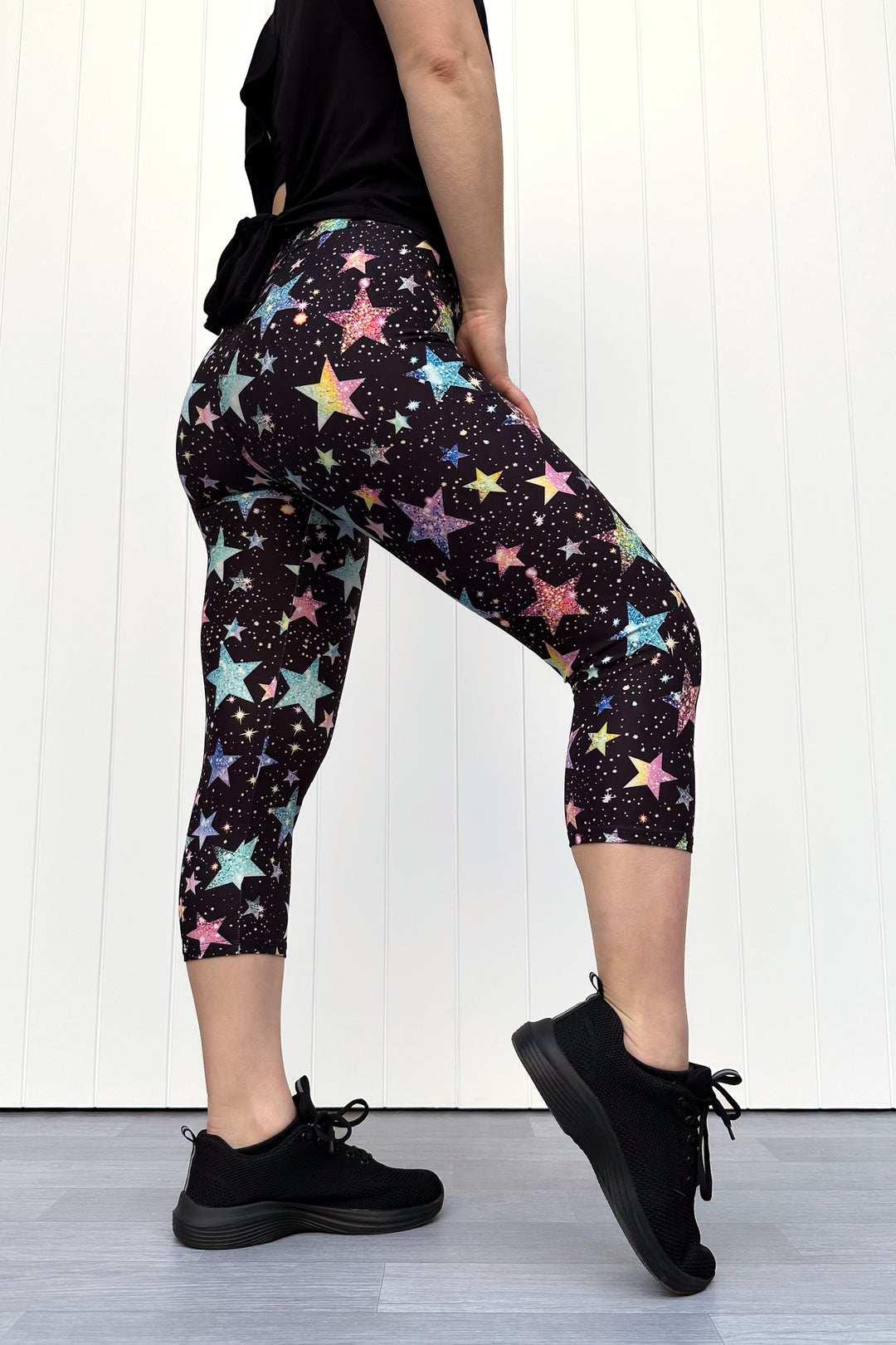 Person wearing black capri leggings with colourful sparkle-like star pattern, standing against a white wall.