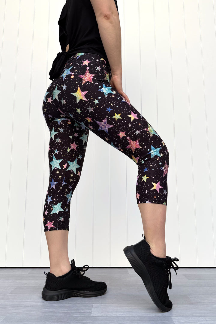 Person wearing black capri leggings with colourful sparkle-like star pattern, standing against a white wall.