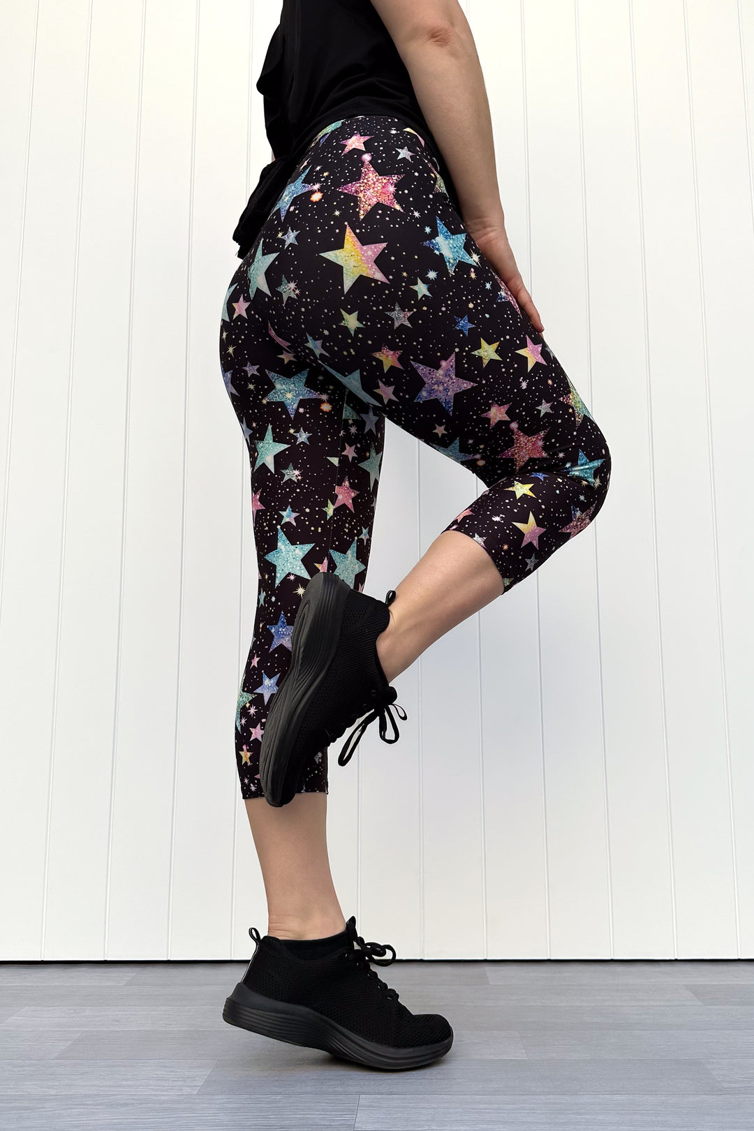 Person wearing black capri leggings with colourful sparkle-like star pattern, standing against a white wall.