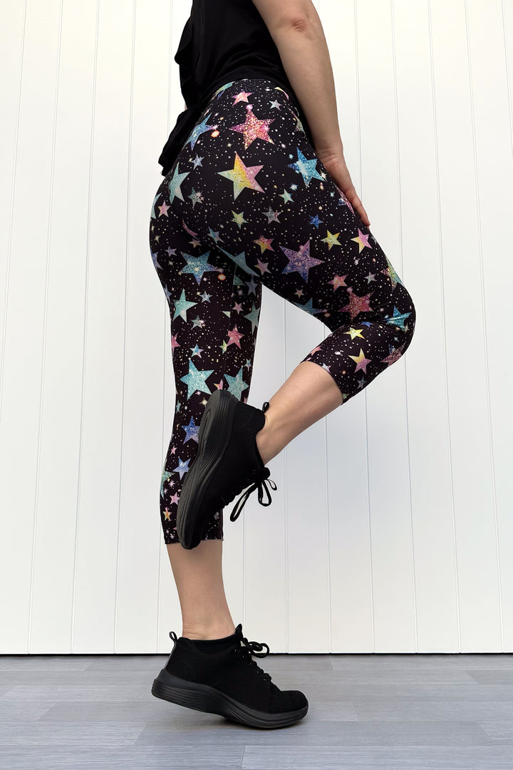 Person wearing black capri leggings with colourful sparkle-like star pattern, standing against a white wall.