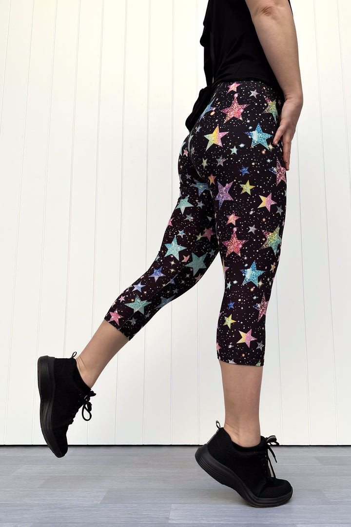 Person wearing black capri leggings with colourful sparkle-like star pattern, standing against a white wall.