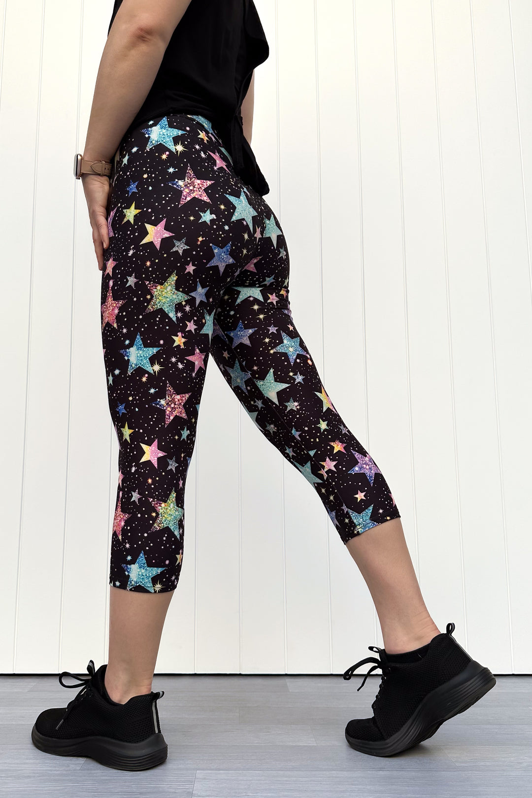 Person wearing black capri leggings with colourful sparkle-like star pattern, standing against a white wall.