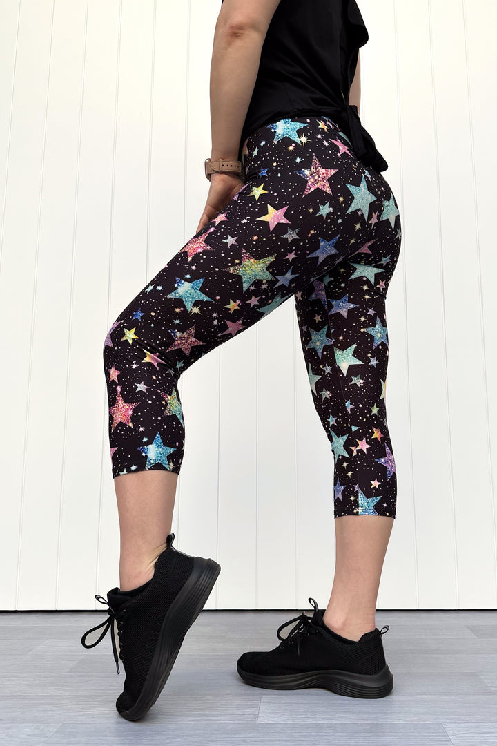 Person wearing black capri leggings with colourful sparkle-like star pattern, standing against a white wall.