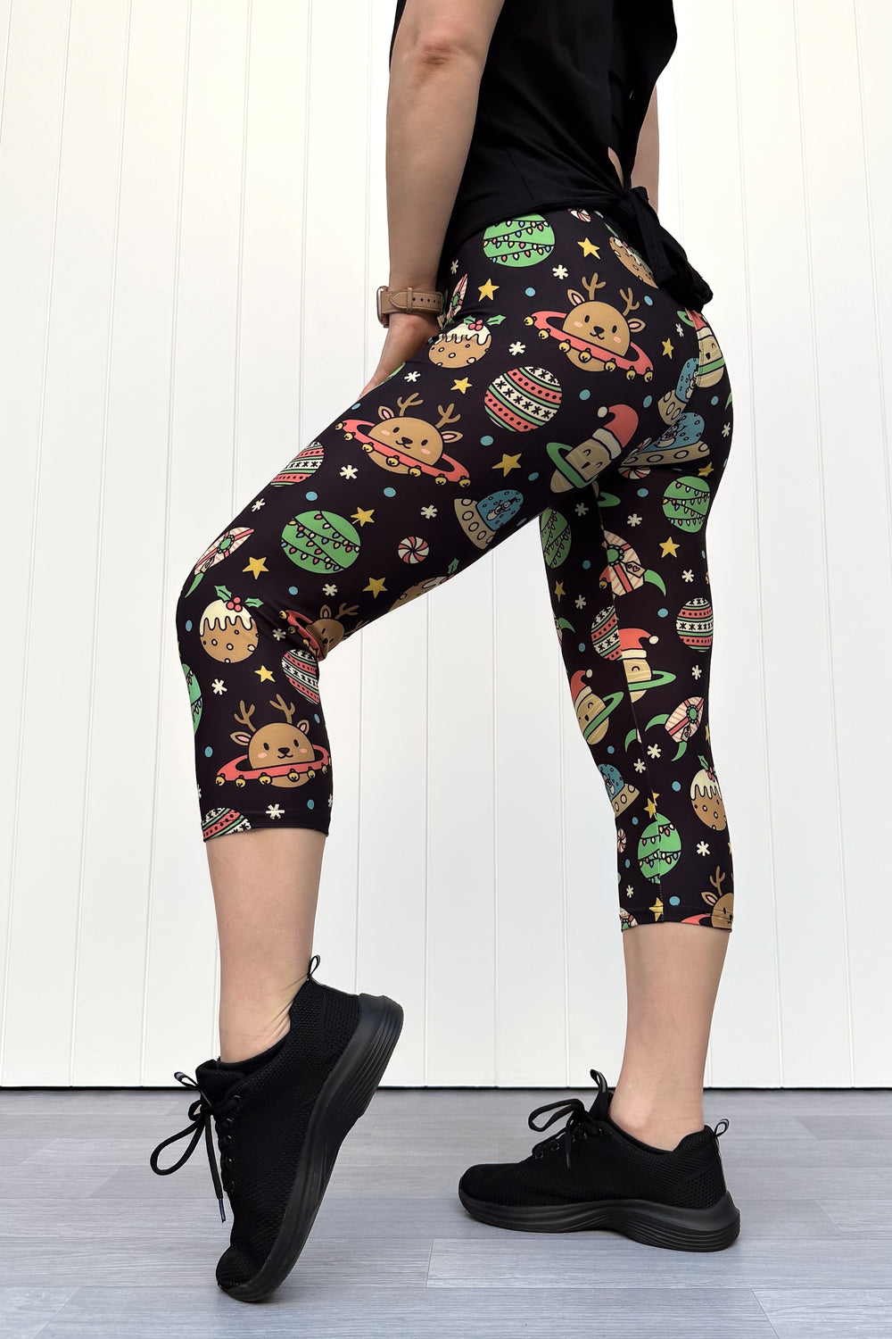 Person wearing black capri leggings with colorful space-themed Christmas pattern standing against a white wall.