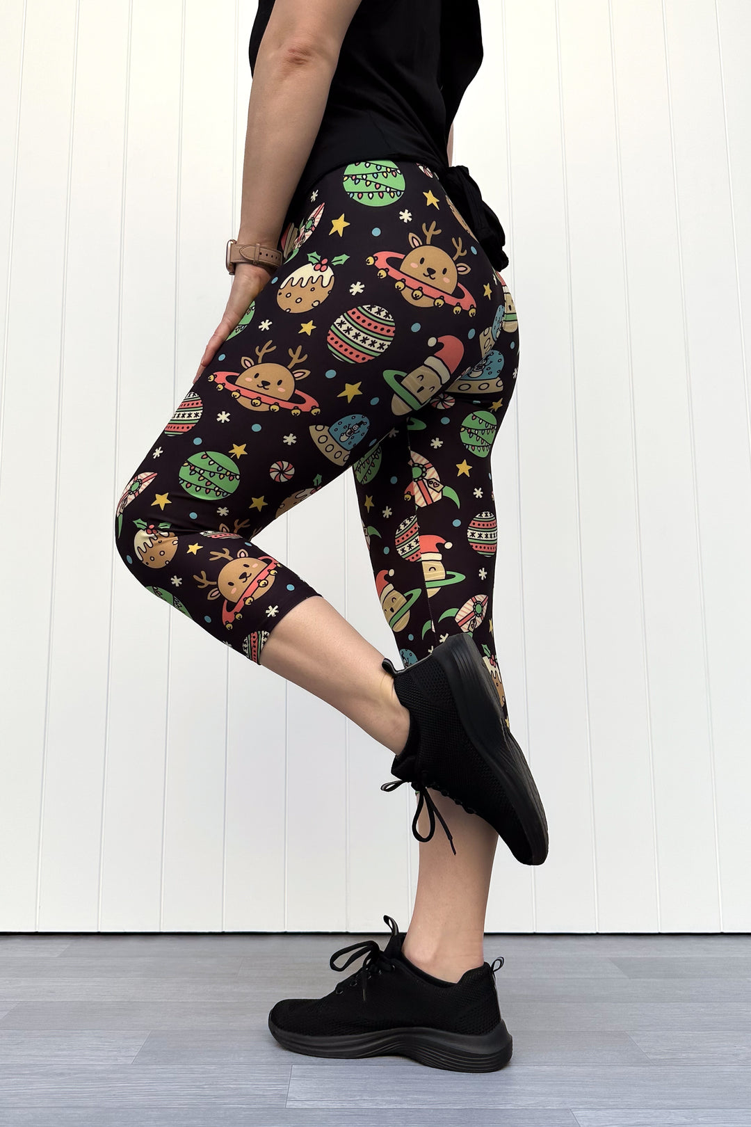 Person wearing black capri leggings with colorful space-themed Christmas pattern standing against a white wall.