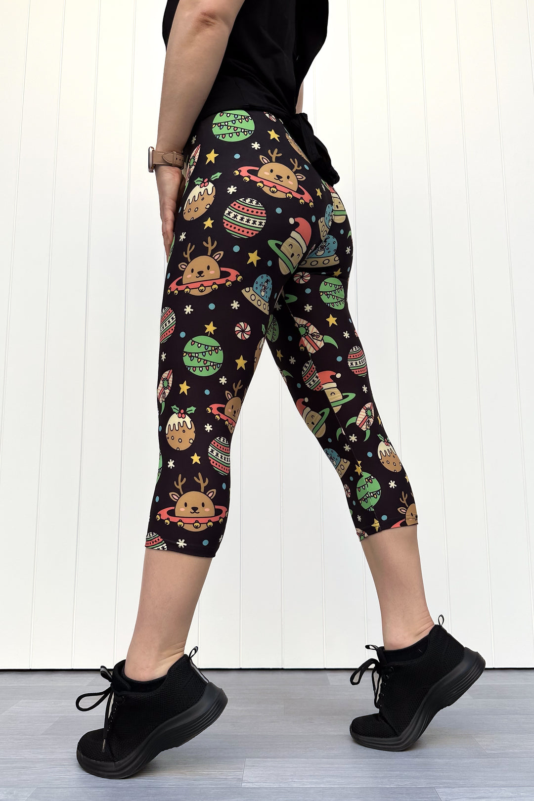 Person wearing black capri leggings with colorful space-themed Christmas pattern standing against a white wall.