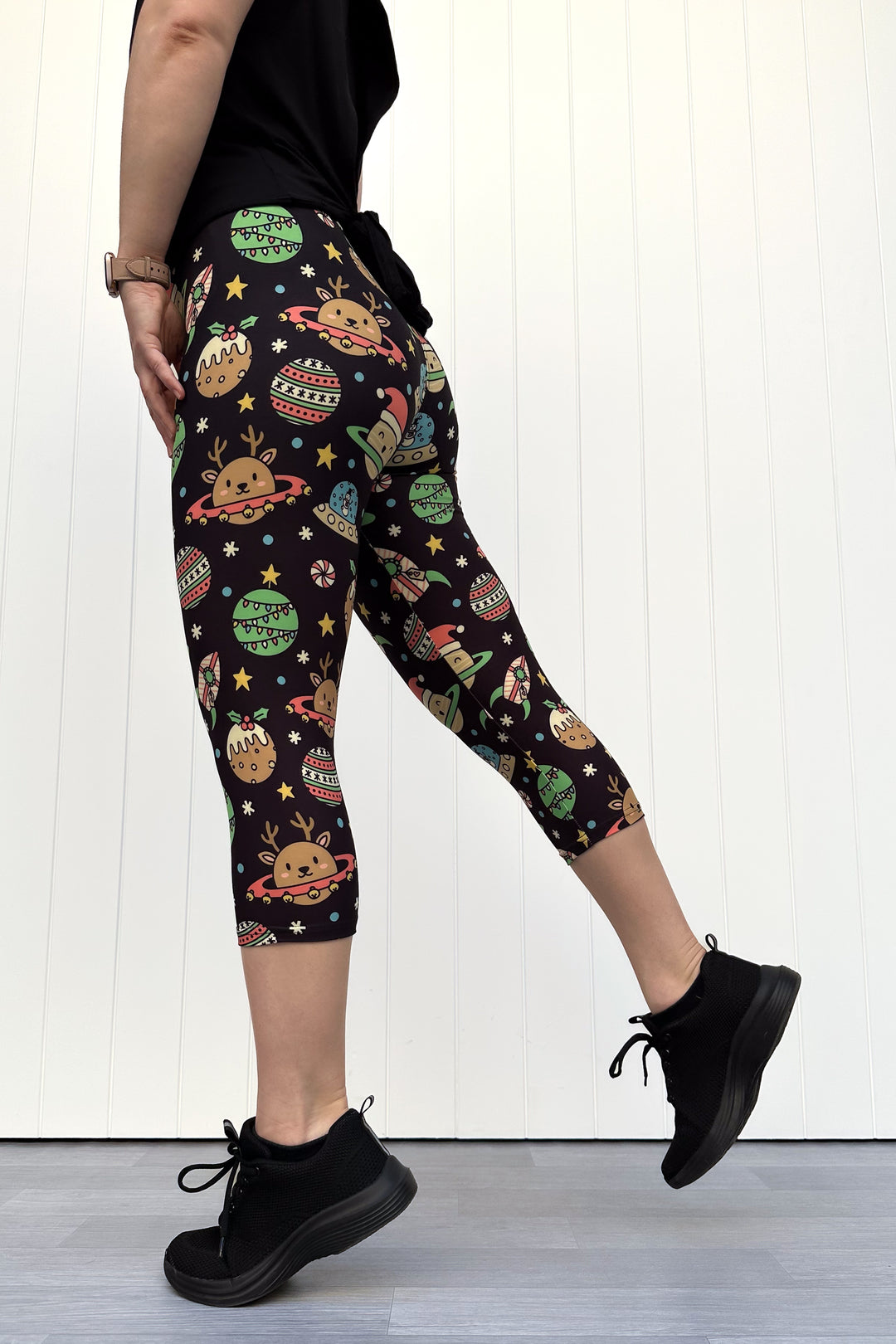Person wearing black capri leggings with colorful space-themed Christmas pattern standing against a white wall.