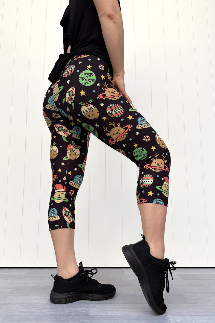 Person wearing black capri leggings with colorful space-themed Christmas pattern standing against a white wall.