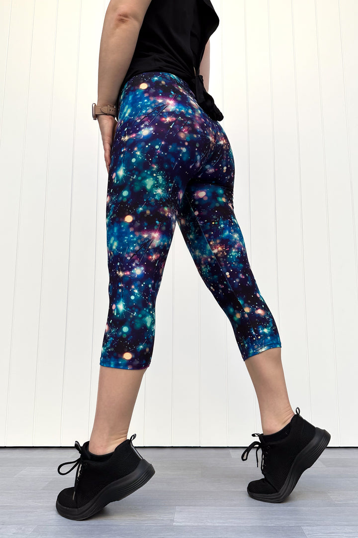 Person wearing celebration sparkler-inspired patterned capri leggings in blues, greens and pinks, standing against a white wall.