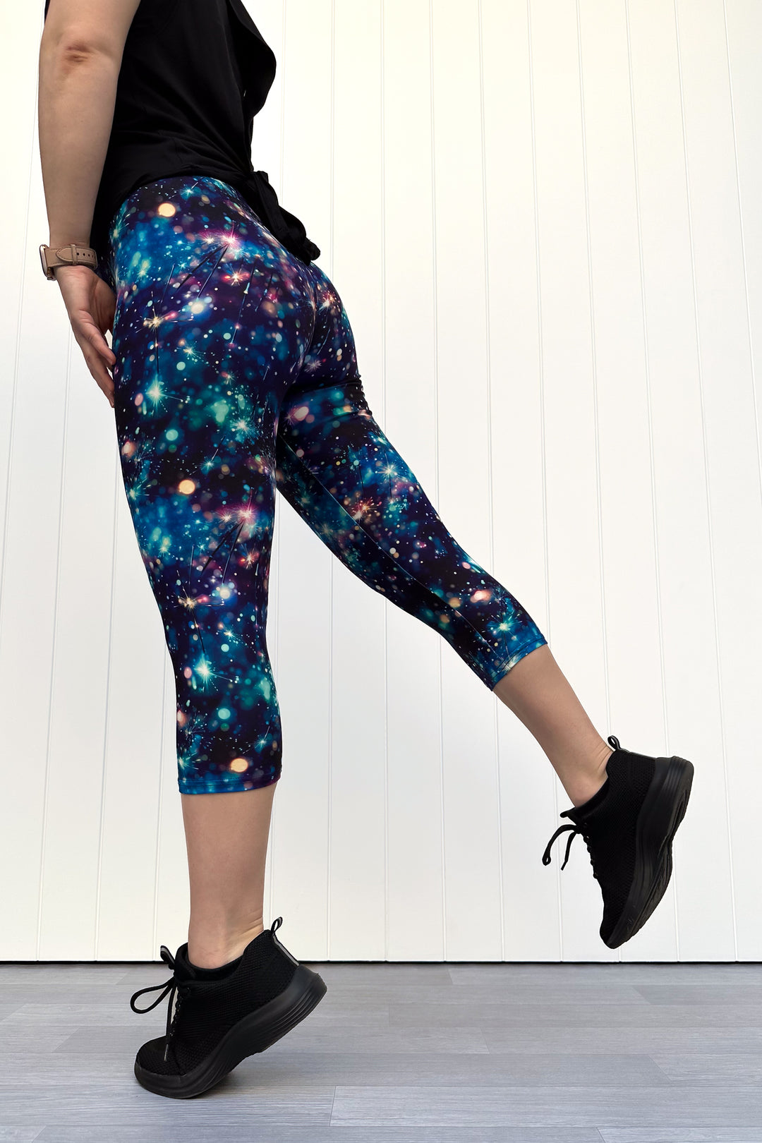 Person wearing celebration sparkler-inspired patterned capri leggings in blues, greens and pinks, standing against a white wall.