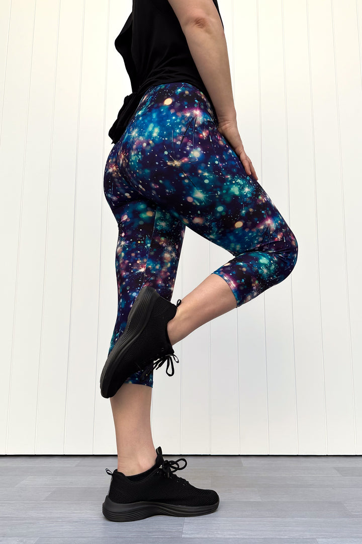 Person wearing celebration sparkler-inspired patterned capri leggings in blues, greens and pinks, standing against a white wall.