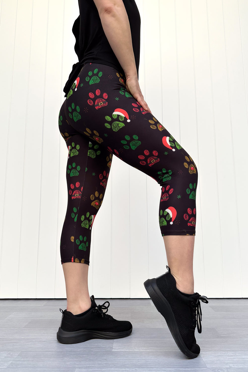 Person wearing black capri leggings with red, green and yellow paw print pattern with santa hats, standing against a white wooden panel background