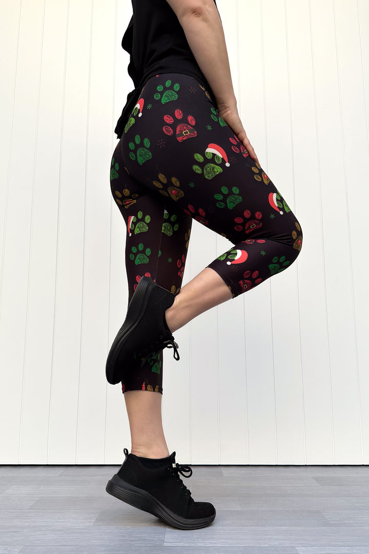 Person wearing black capri leggings with red, green and yellow paw print pattern with santa hats, standing against a white wooden panel background
