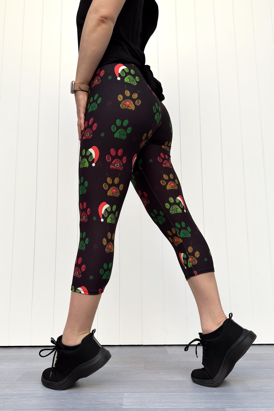 Person wearing black capri leggings with red, green and yellow paw print pattern with santa hats, standing against a white wooden panel background