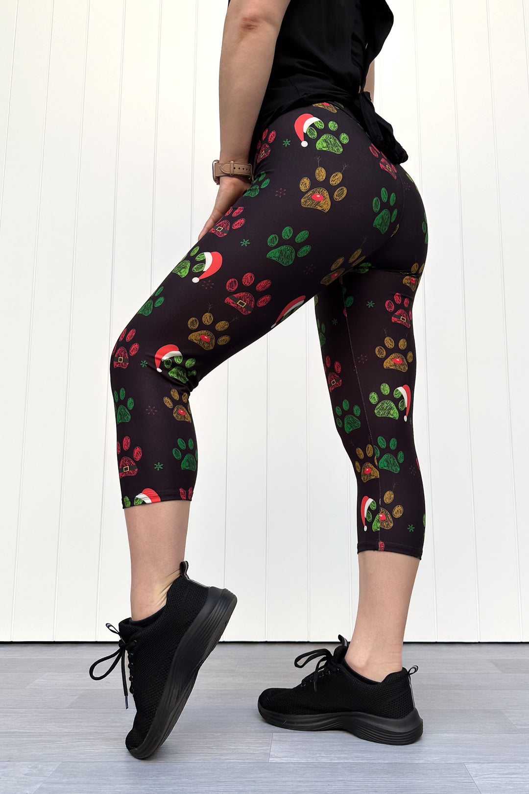 Person wearing black capri leggings with red, green and yellow paw print pattern with santa hats, standing against a white wooden panel background