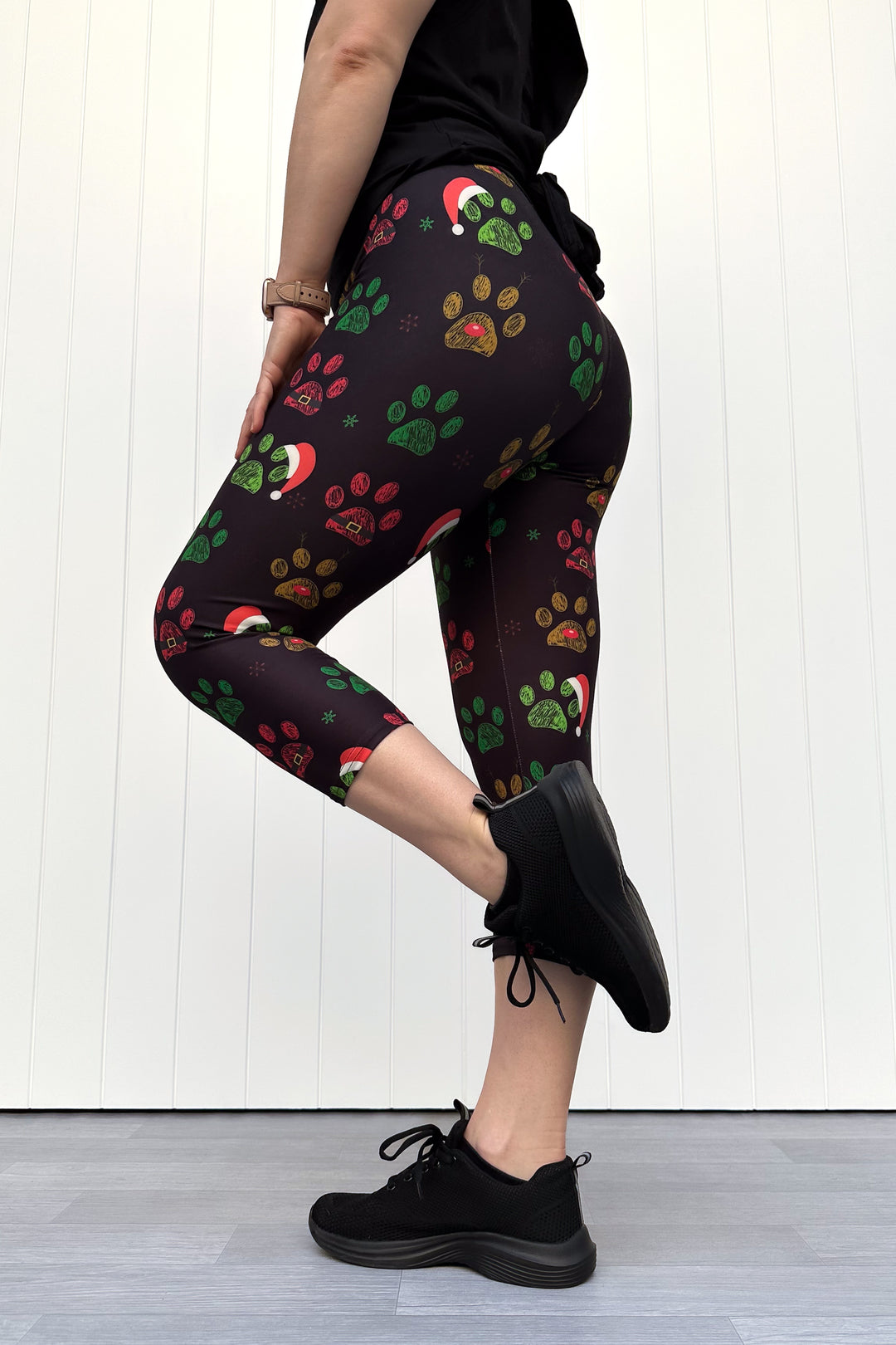 Person wearing black capri leggings with red, green and yellow paw print pattern with santa hats, standing against a white wooden panel background