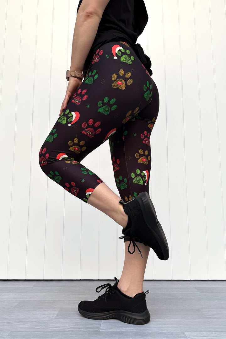 Person wearing black capri leggings with red, green and yellow paw print pattern with santa hats, standing against a white wooden panel background