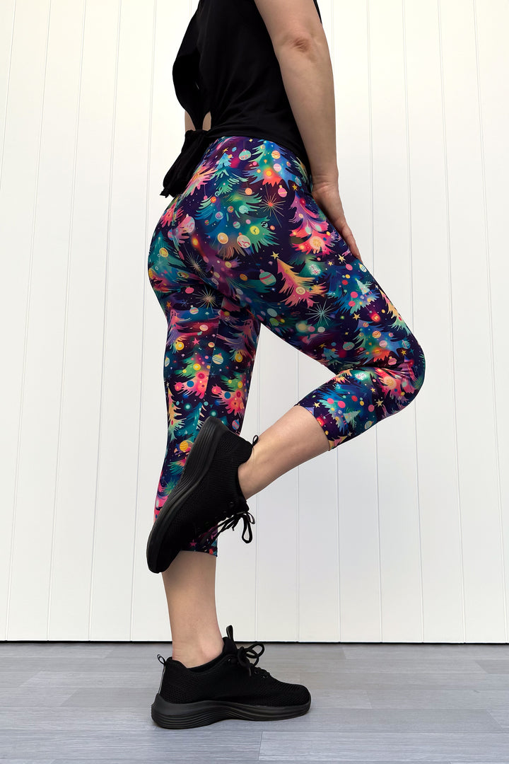 Person wearing capri leggings with a pattern of colourful Christmas trees against a white wooden panel background.