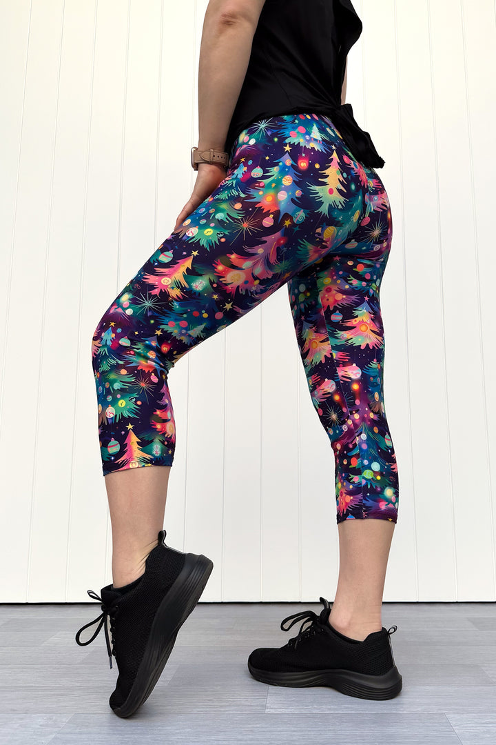 Person wearing capri leggings with a pattern of colourful Christmas trees against a white wooden panel background.