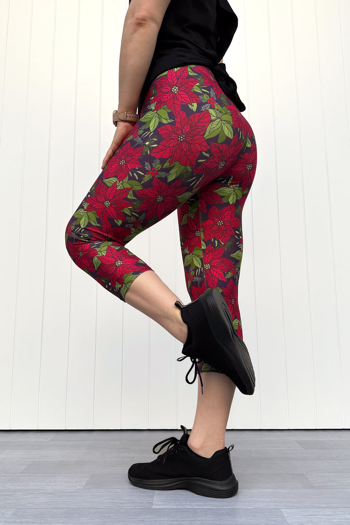 Person wearing red floral Christmas capri leggings, standing against a white wall.