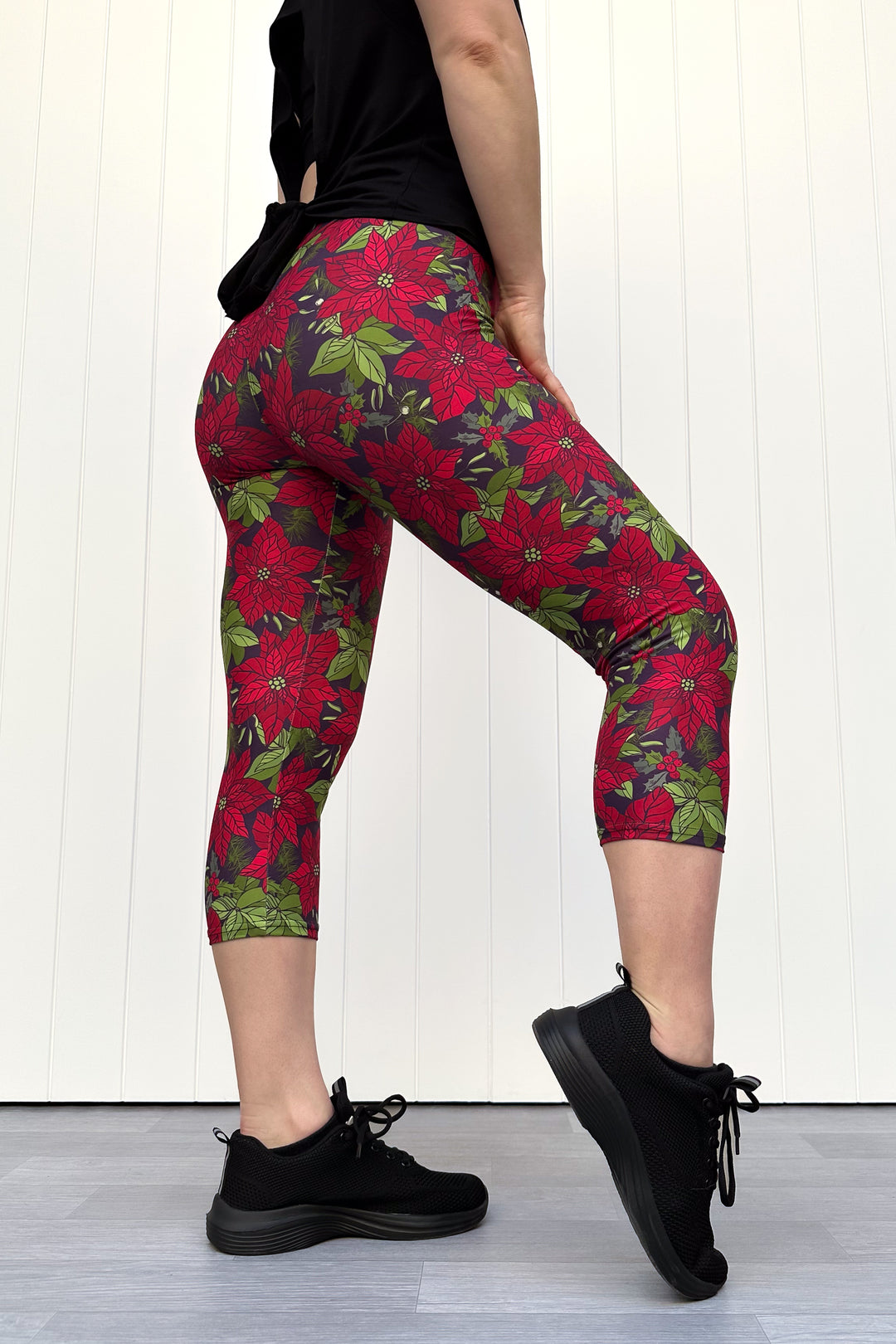 Person wearing red floral Christmas capri leggings, standing against a white wall.
