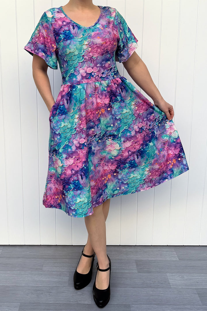 Person wearing a colourful mermaid fantasy pattern dress against a white wooden panel background