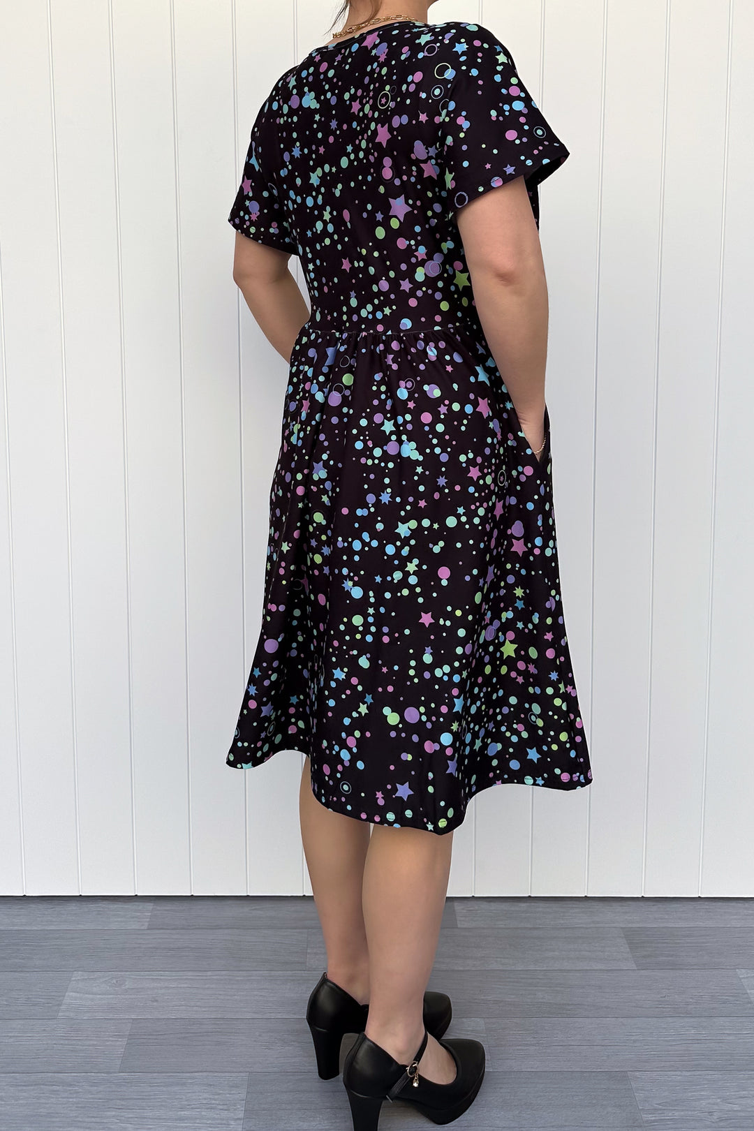 Person wearing a black dress with colorful patterns against a white wooden panel background