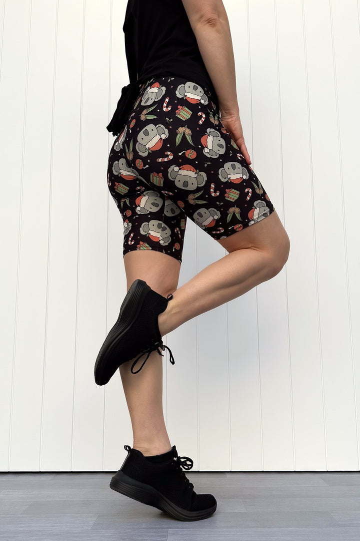 Person wearing shorts featuring a pattern of koalas wearing santa hats and candy canes on a black background, standing against a white wall.
