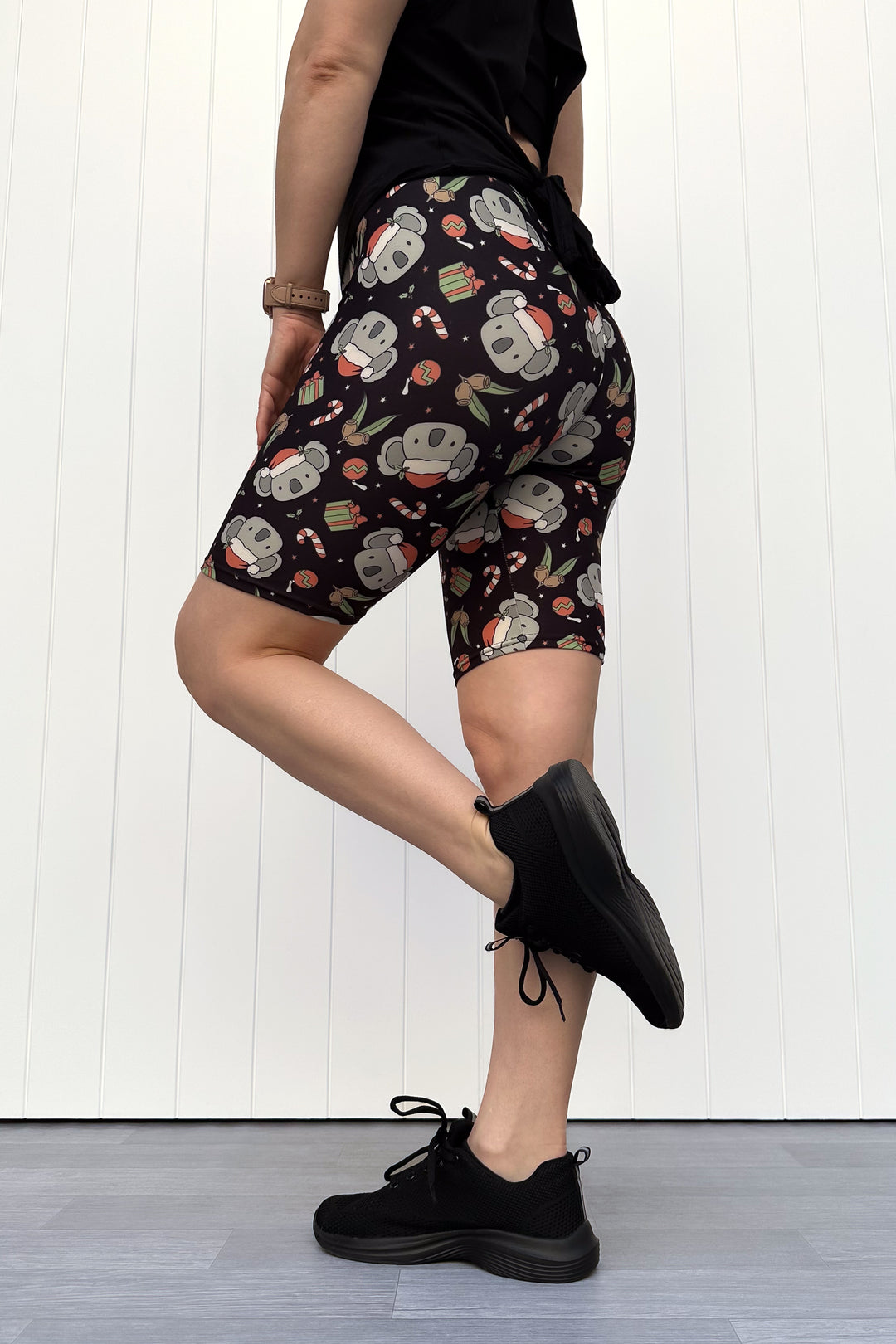 Person wearing shorts featuring a pattern of koalas wearing santa hats and candy canes on a black background, standing against a white wall.