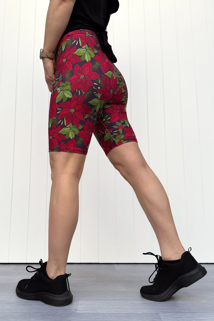 Person wearing red floral Christmas shorts, standing against a white wall.