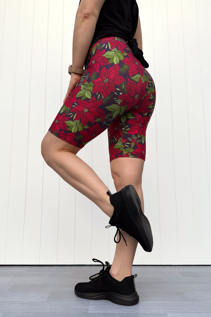 Person wearing red floral Christmas shorts, standing against a white wall.