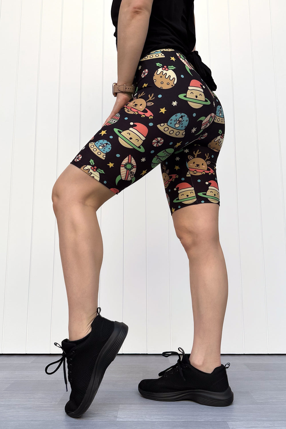 Person wearing black shorts with colorful space-themed Christmas pattern standing against a white wall.