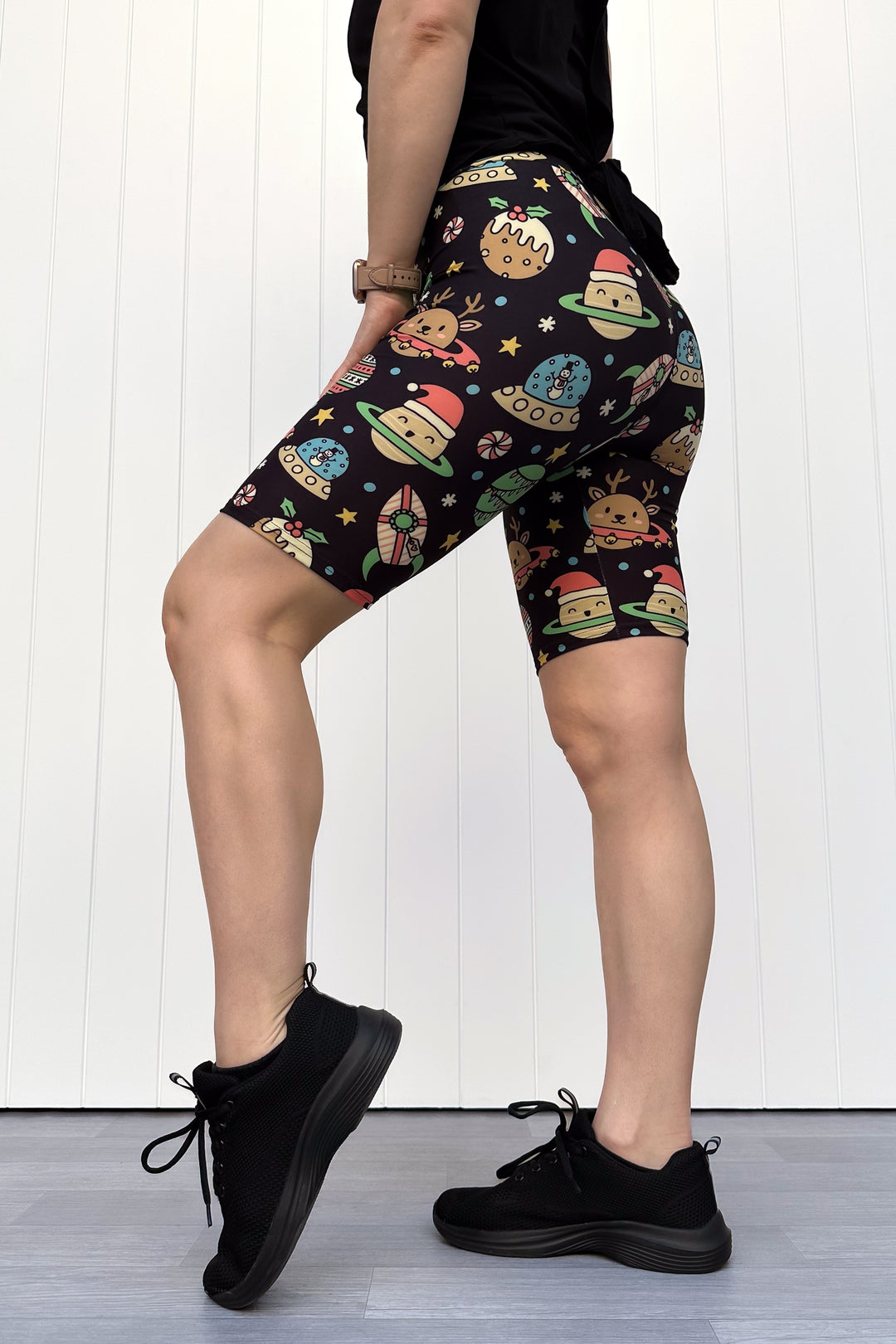 Person wearing black shorts with colorful space-themed Christmas pattern standing against a white wall.