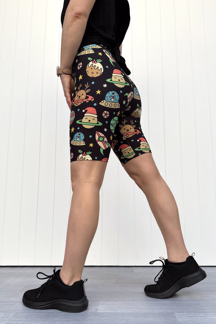 Person wearing black shorts with colorful space-themed Christmas pattern standing against a white wall.