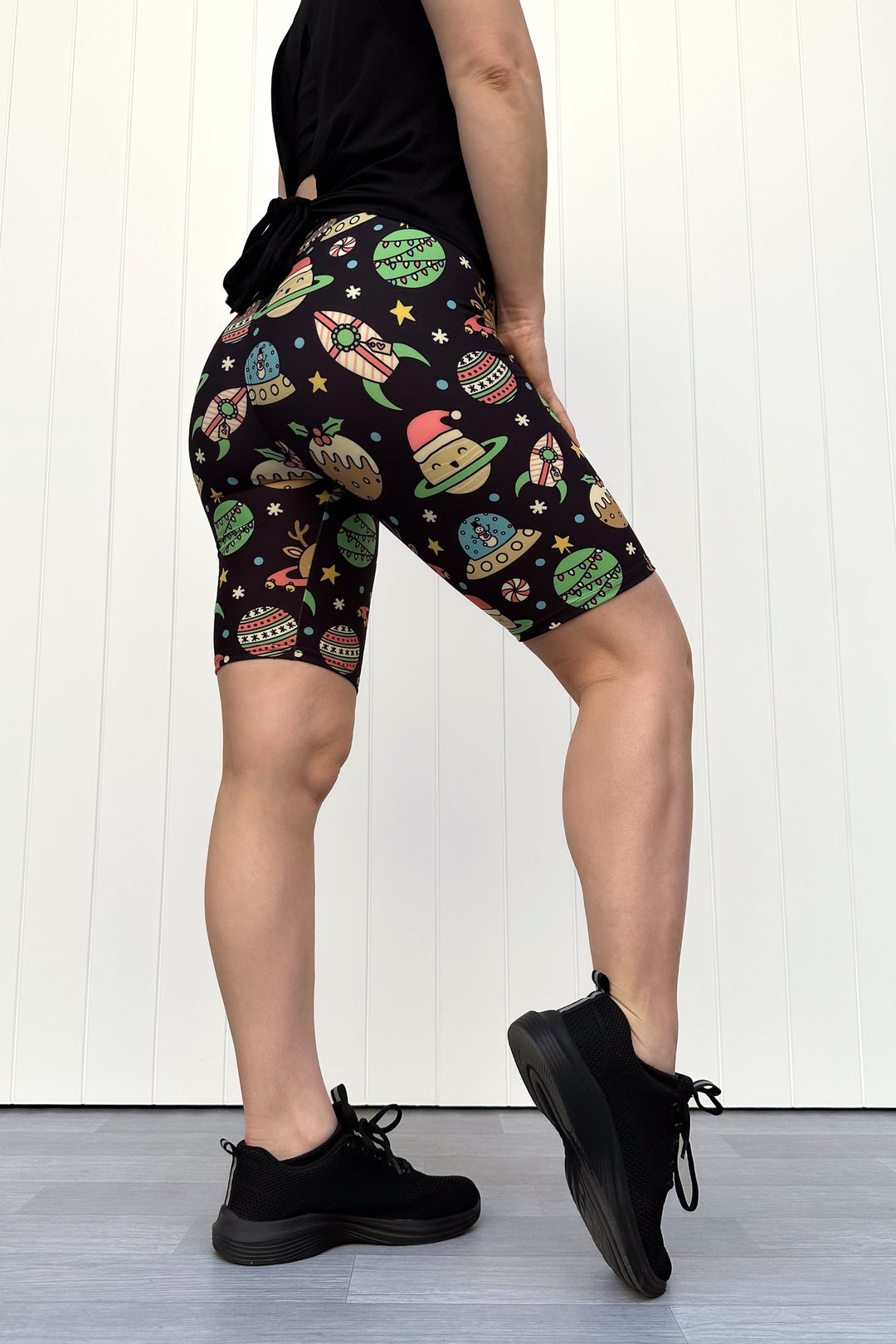 Person wearing black shorts with colorful space-themed Christmas pattern standing against a white wall.