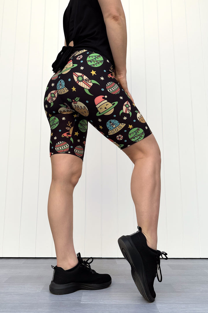 Person wearing black shorts with colorful space-themed Christmas pattern standing against a white wall.