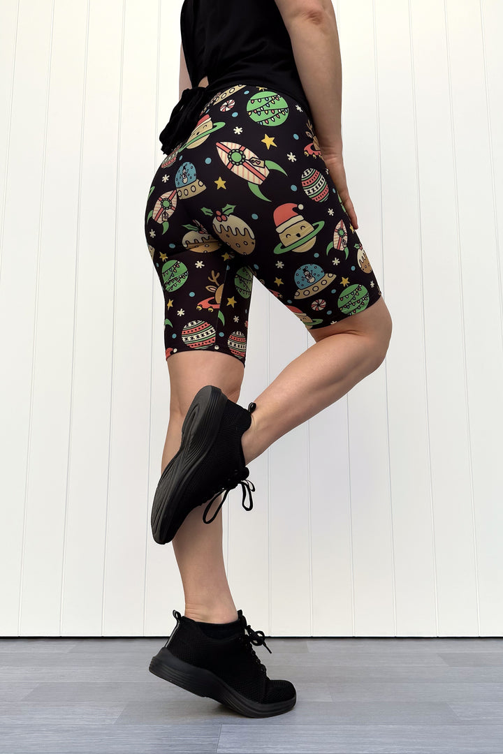 Person wearing black shorts with colorful space-themed Christmas pattern standing against a white wall.