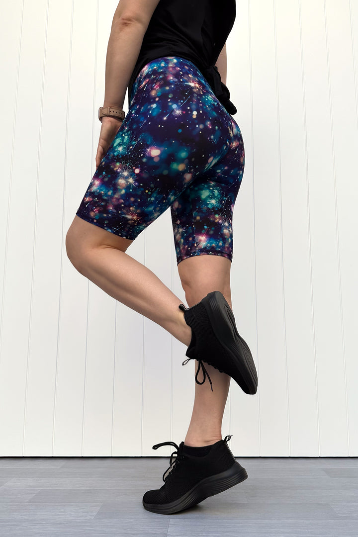 Person wearing celebration sparkler-inspired patterned shorts in blues, greens and pinks, standing against a white wall.