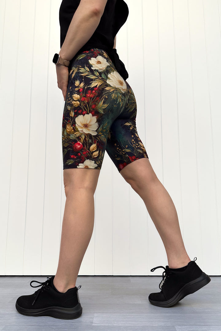 Person wearing dark Christmas-themed floral shorts with white flowers and red berries, standing against a white wooden panel background