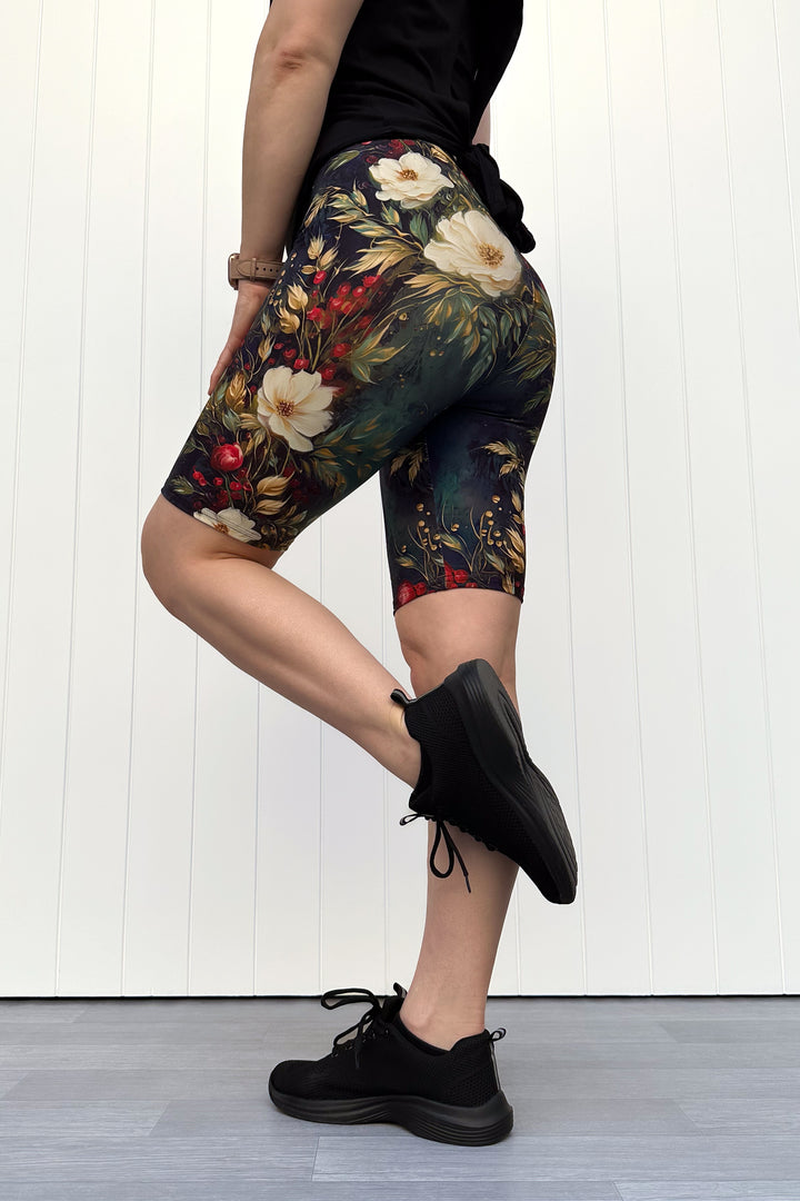 Person wearing dark Christmas-themed floral shorts with white flowers and red berries, standing against a white wooden panel background