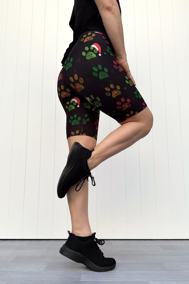 Person wearing black shorts with red, green and yellow paw print pattern with santa hats, standing against a white wooden panel background
