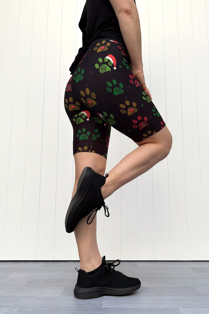 Person wearing black shorts with red, green and yellow paw print pattern with santa hats, standing against a white wooden panel background