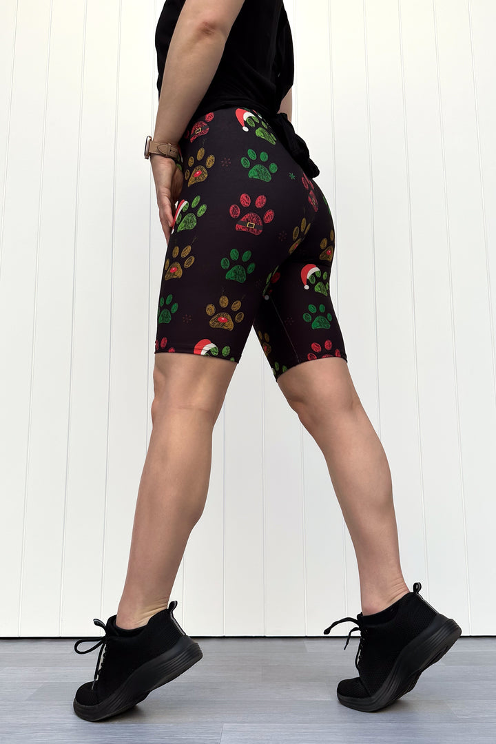 Person wearing black shorts with red, green and yellow paw print pattern with santa hats, standing against a white wooden panel background