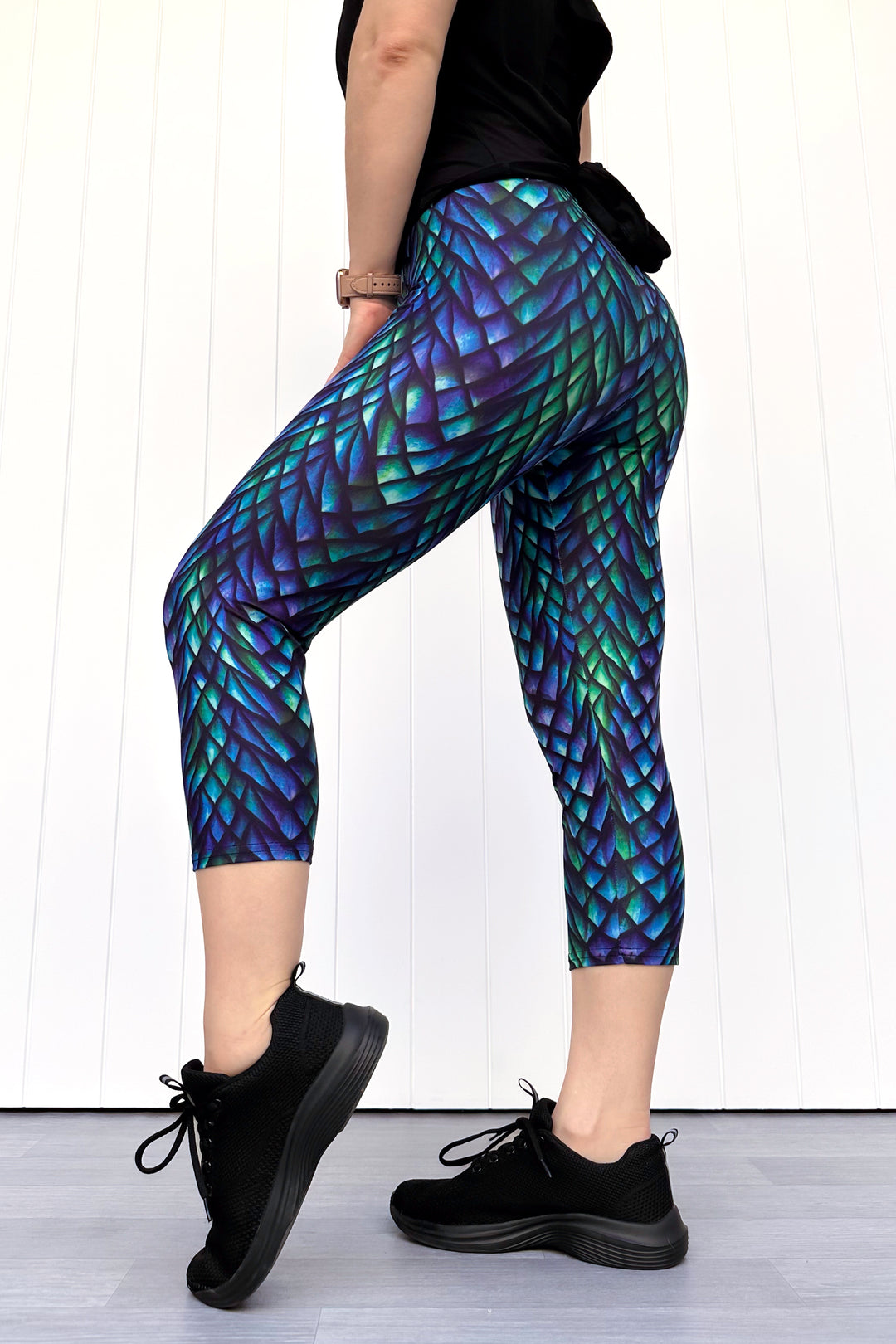 Person wearing green and purple capri leggings with a dragon scale pattern, standing against a white wall.