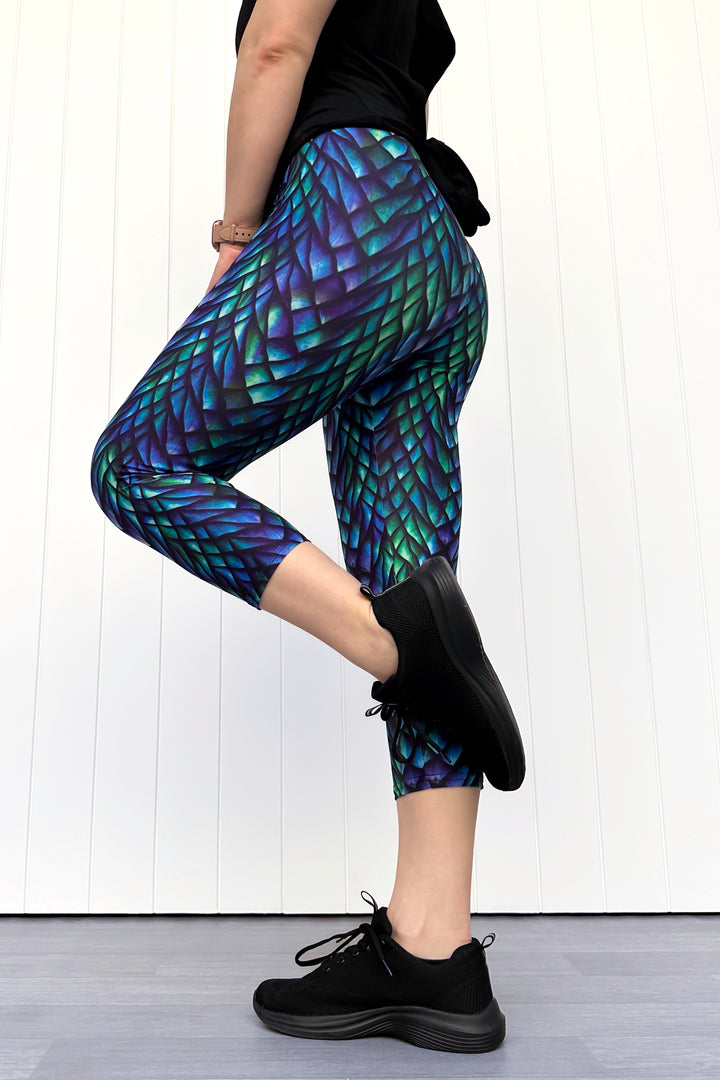 Person wearing green and blue capri leggings with a dragon scale pattern, standing against a white wall.