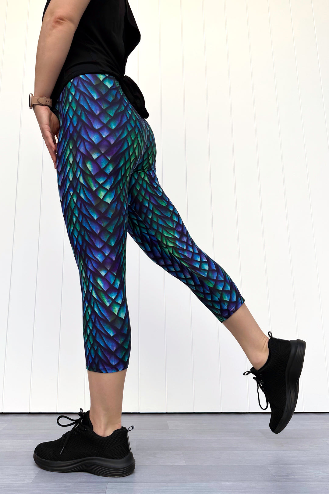 Person wearing green and purple capri leggings with a dragon scale pattern, standing against a white wall.
