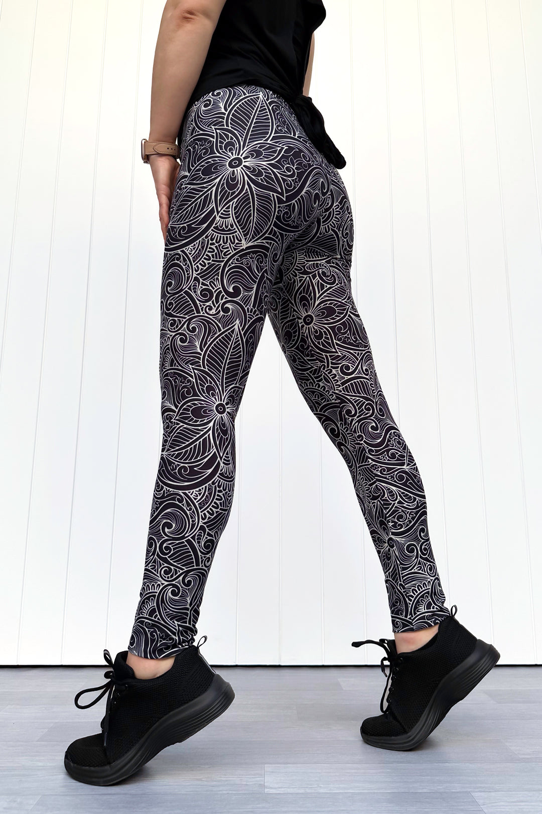 Person wearing black patterned leggings with black shoes on a light wooden floor.