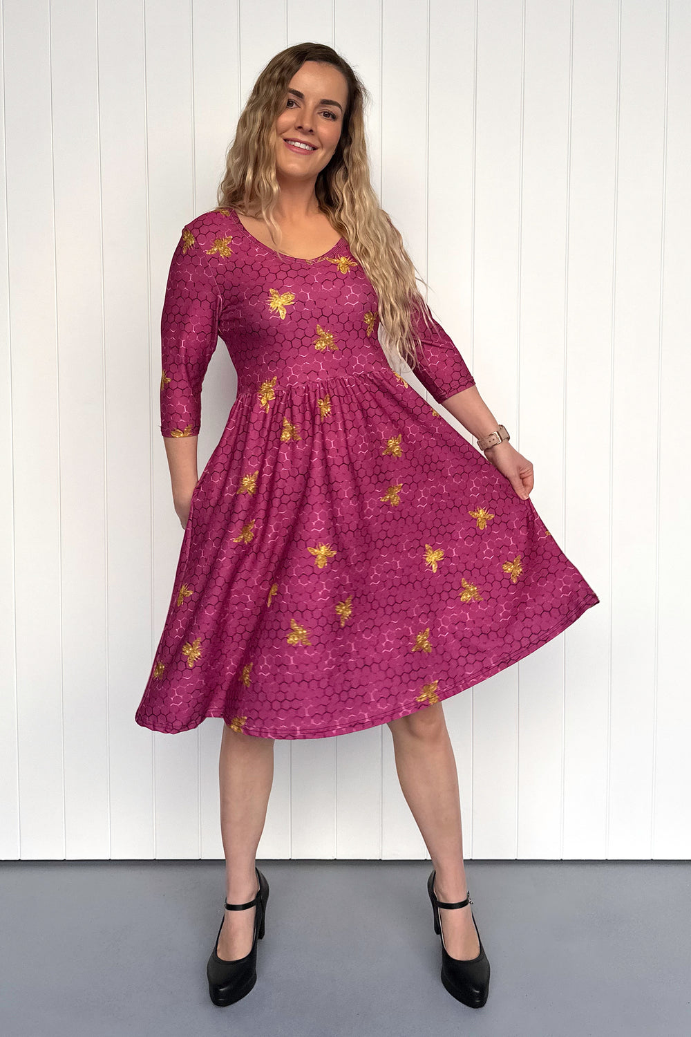 Woman wearing a magenta dress with golden bee patterns against a white wall.